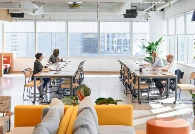 Bright open-plan coworking area with long wooden tables and vibrant orange chairs.