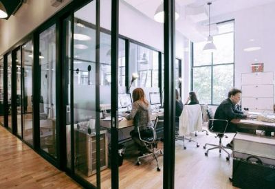 Sunlit open-plan office with glass partitions and several wooden desks with ergonomic chairs.