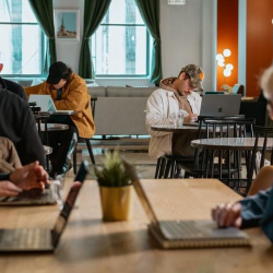 Bright open-plan workspace with people working at communal wooden tables.