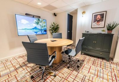 Round meeting table with blue chairs, a wall-mounted screen, and patterned carpet.