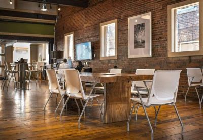 Bright communal workspace with a large rustic wooden table, white chairs, and exposed brick walls.