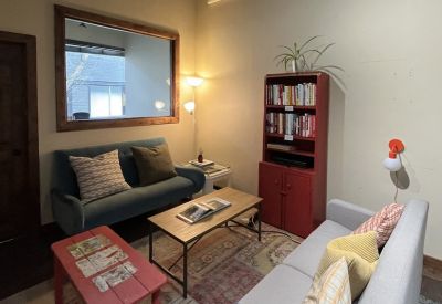Bright communal lounge with colorful rugs, plush sofas, a red bookcase, and a large internal window.