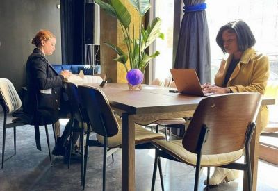 Two people working at a wooden table in a sunlit room with potted plants.