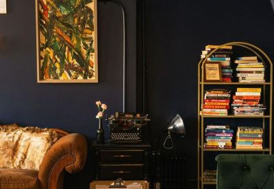 Vintage lounge nook with a brown velvet sofa, antique typewriter, and a gold bookshelf against a dark wall.