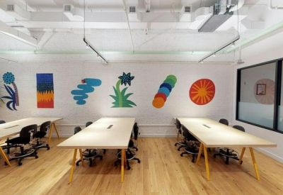Modern open-plan office workspace with long light-wood desks and colorful wall illustrations.