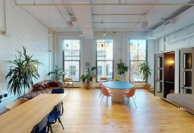 Open breakout area with a blue round table, orange chairs, and indoor plants by large windows.