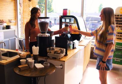 Bright reception and cafe counter where a barista serves a customer near a coffee grinder.