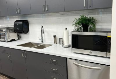 Modern office breakroom with grey cabinetry, white countertops, and stainless steel appliances.