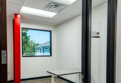 Private office suite with a white desk, grey carpet, and a view through a large window.