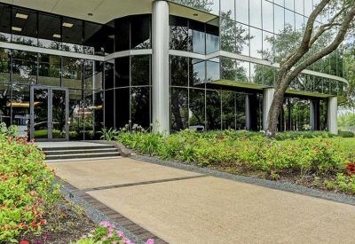 Modern glass building entrance with landscaped flower beds and trees.