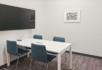 A small meeting room with a white table, teal chairs, and a large wall-mounted monitor.