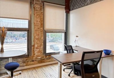 Bright workspace with an L-shaped desk, ergonomic chairs, and brick accents by large windows.