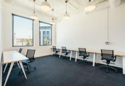 Bright open-plan office space with shared wooden desks and black ergonomic chairs.