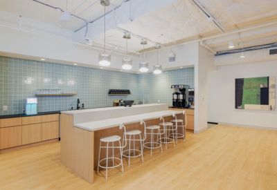Modern office kitchen area with a light wood island, white stools, and teal tile backsplash.