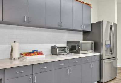 Modern office kitchen featuring grey cabinetry, white countertops, and a stainless steel refrigerator.