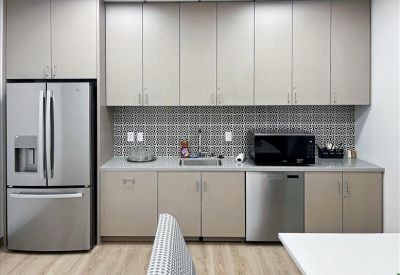 Modern office kitchen with tan cabinets, grey tile backsplash, and stainless steel appliances.