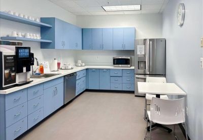 Bright office kitchen with light blue cabinetry, stainless steel fridge, and a small dining table.