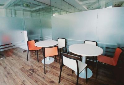 Bright communal break area with white round tables, orange accent chairs, and frosted glass partitions.