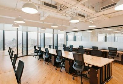 Long open-plan office space featuring wooden desks, black ergonomic chairs, and large perimeter windows.