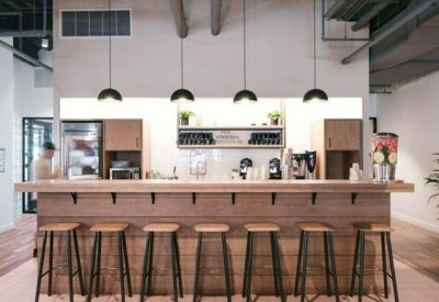 Modern office kitchen and cafe area featuring a wooden bar with several stools and pendant lighting.