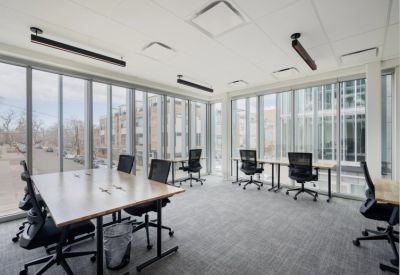 Bright corner office with multiple desks, black mesh chairs, and floor-to-ceiling windows.