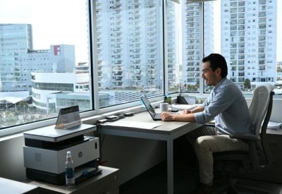 A man working at a bright desk by large windows with cityscape views.