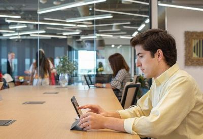Modern open-plan workspace with a person using a tablet at a long light-wood table.