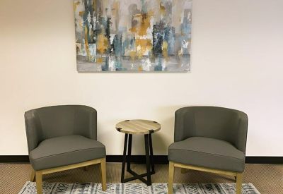 Small seating area with two grey club chairs, a round wooden side table, and abstract wall art.