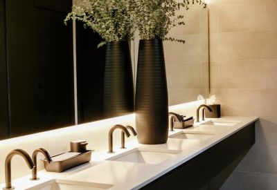 Modern bathroom with a long white vanity, multiple sinks, black fixtures, and large vases of greenery.