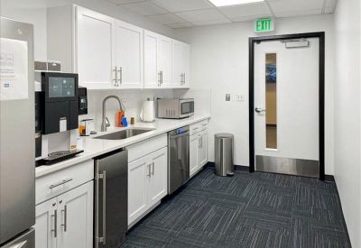 Modern office kitchen with white cabinetry, stainless steel appliances, and dark patterned carpet.
