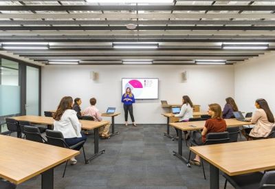 Professional training room with rows of wooden tables, chairs, and a presenter at a digital screen.
