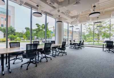 Bright open-plan coworking space featuring long wooden desks and black ergonomic chairs.