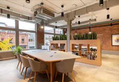Communal wooden dining table with grey chairs near a brick wall and indoor plants.