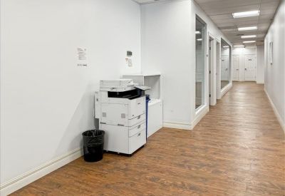 Office corridor with wooden flooring, white walls, and a large multi-function printer.