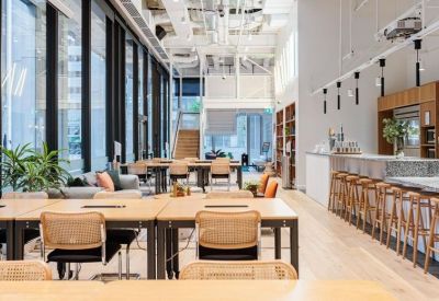 Spacious open-plan coworking area with rows of wooden desks and large glass windows.