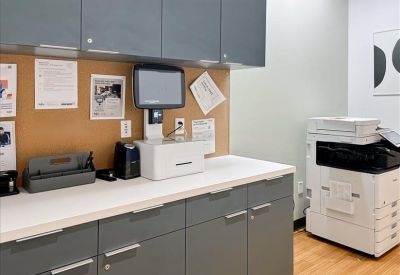 An office print and mail station with grey cabinetry, a corkboard, and a large multifunction printer.