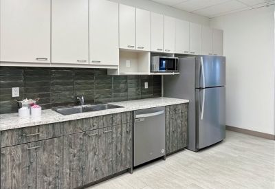 Modern office kitchen with wood-grain cabinets, stainless steel appliances, and a gray backsplash.