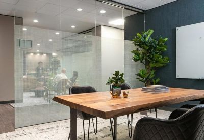 Small meeting room with a wooden table, black chairs, and large indoor plants by a glass wall.