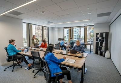 Bright coworking space with professionals working at long wooden tables near large windows.