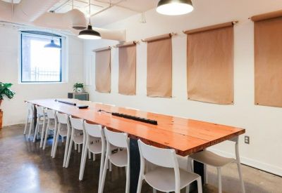 Long communal wooden table with white chairs and brown window shades.
