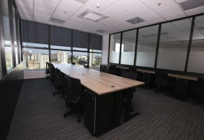Open-plan office space with long wooden desks, black chairs, and large windows with city views.
