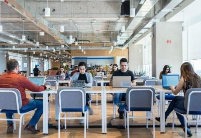 Open-plan coworking area with people working at long white tables under an industrial ceiling.