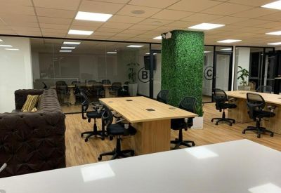 Open-plan coworking space with light wood desks, ergonomic chairs, and a green moss feature pillar.