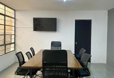 Bright conference room with a long wooden table, black mesh chairs, and a wall-mounted TV.