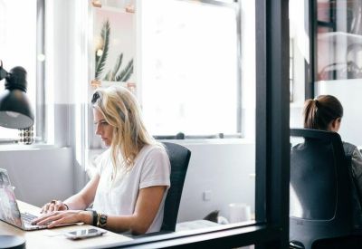 Bright workspace with a person working at a desk beside a large window and glass partitions.