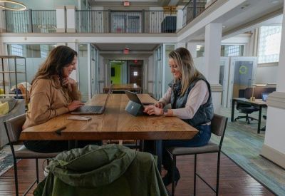 Coworkers collaborating at a large natural wood communal table in a bright open-plan space.