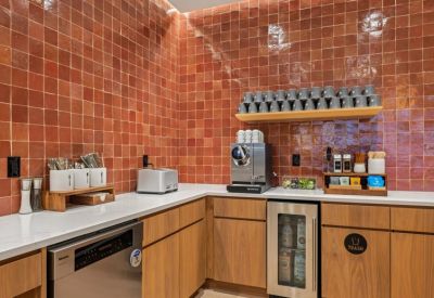 Modern communal kitchen with red tile backsplash, espresso machine, and light wood cabinetry.