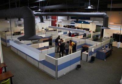 Large-scale office layout with numerous cubicles and exposed dark ceiling ventilation ducts.