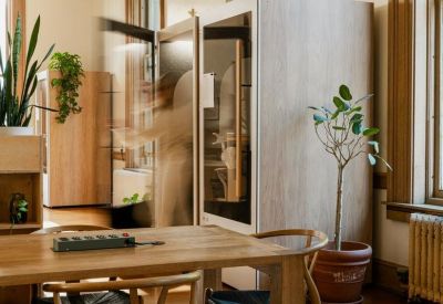 Portrait view of a bright workspace with a privacy phone booth, wooden table, and lush green plants.