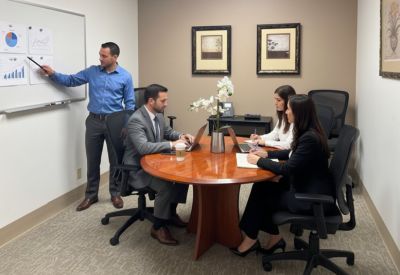 Four professionals collaborating around a round wood table with a whiteboard in the background.